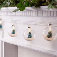 An einem Kamin aufgespannte Lichterkette mit Weihnachtsbaumkugeln aus Glas mit jeweils einem Mini-Weihnachtsbaum und Kunstschnee im Inneren