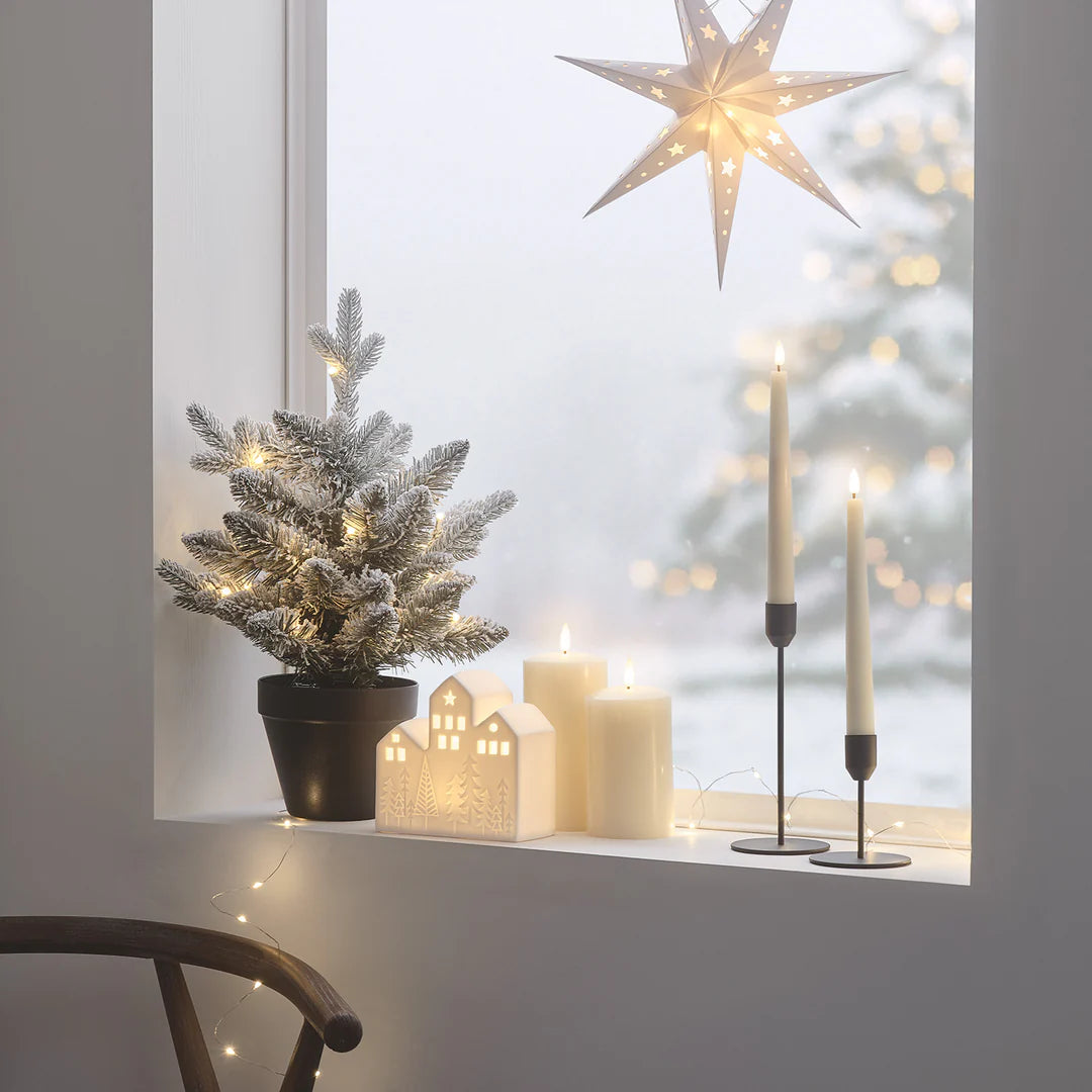Beleuchteter verschneiter LED-Weihnachtsbaum auf Fensterbank mit Kerzen und Papierstern