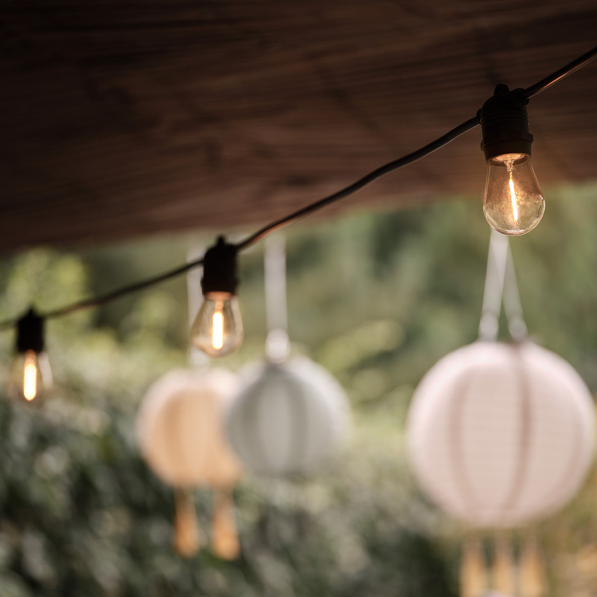 Nahaufnahme einer leuchtenden Glühbirne an schwarzem Kabel mit Papierlaternen im Hintergrund