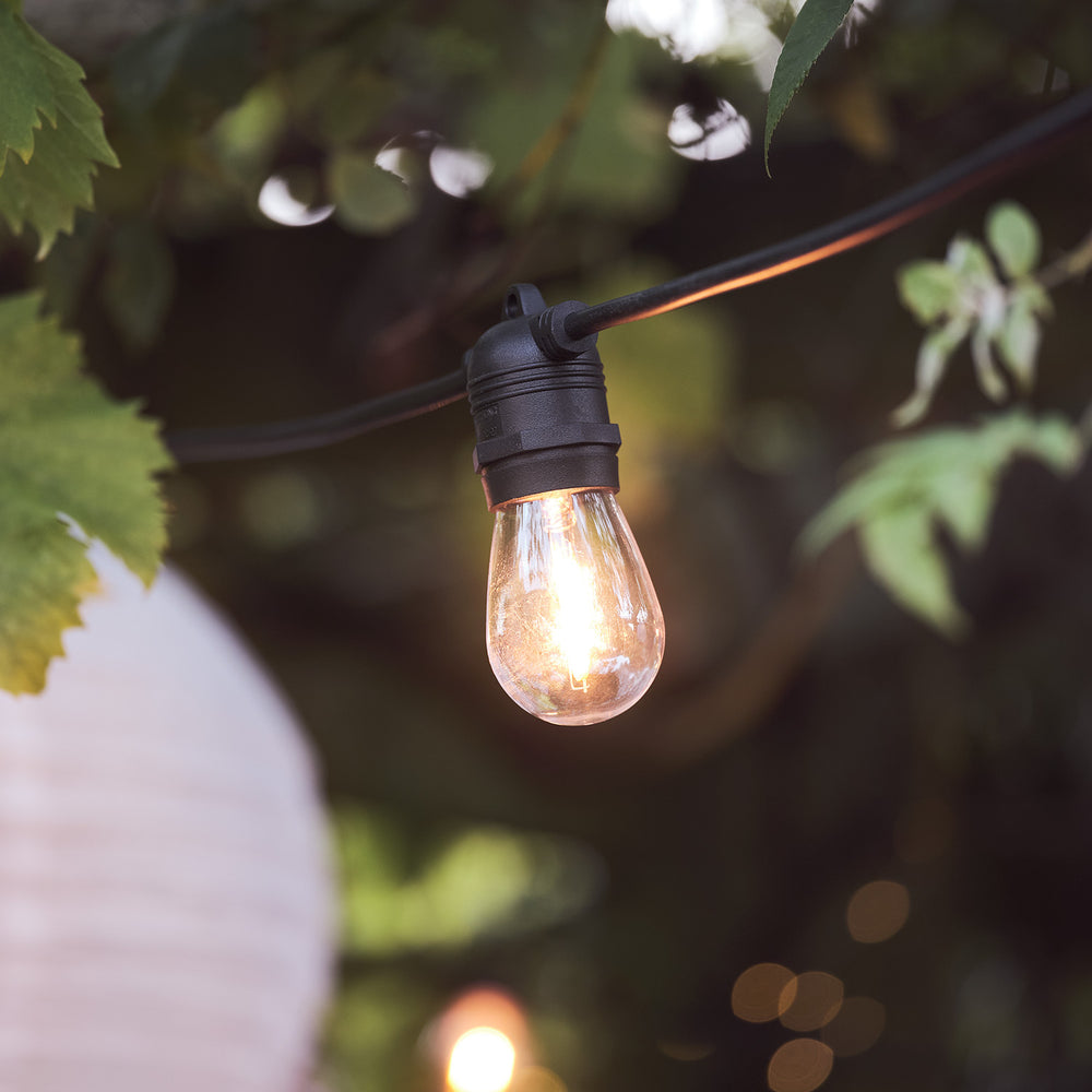 Nahaufnahme einer warmweißen Glühbirne an schwarzem Kabel im Garten