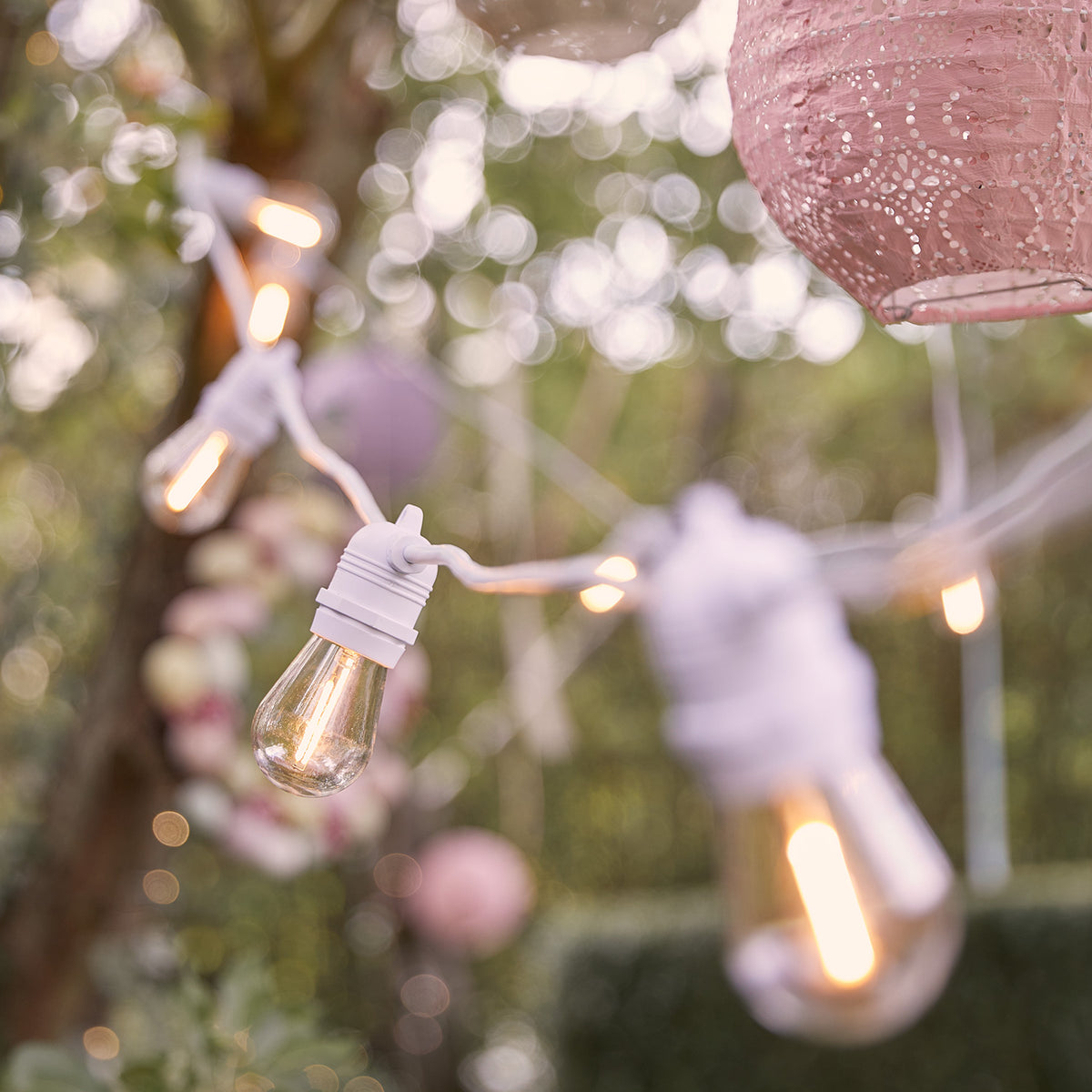 Lichterkette mit warmweißen Glühbirnen an weißem Kabel im Garten mit Dekoration