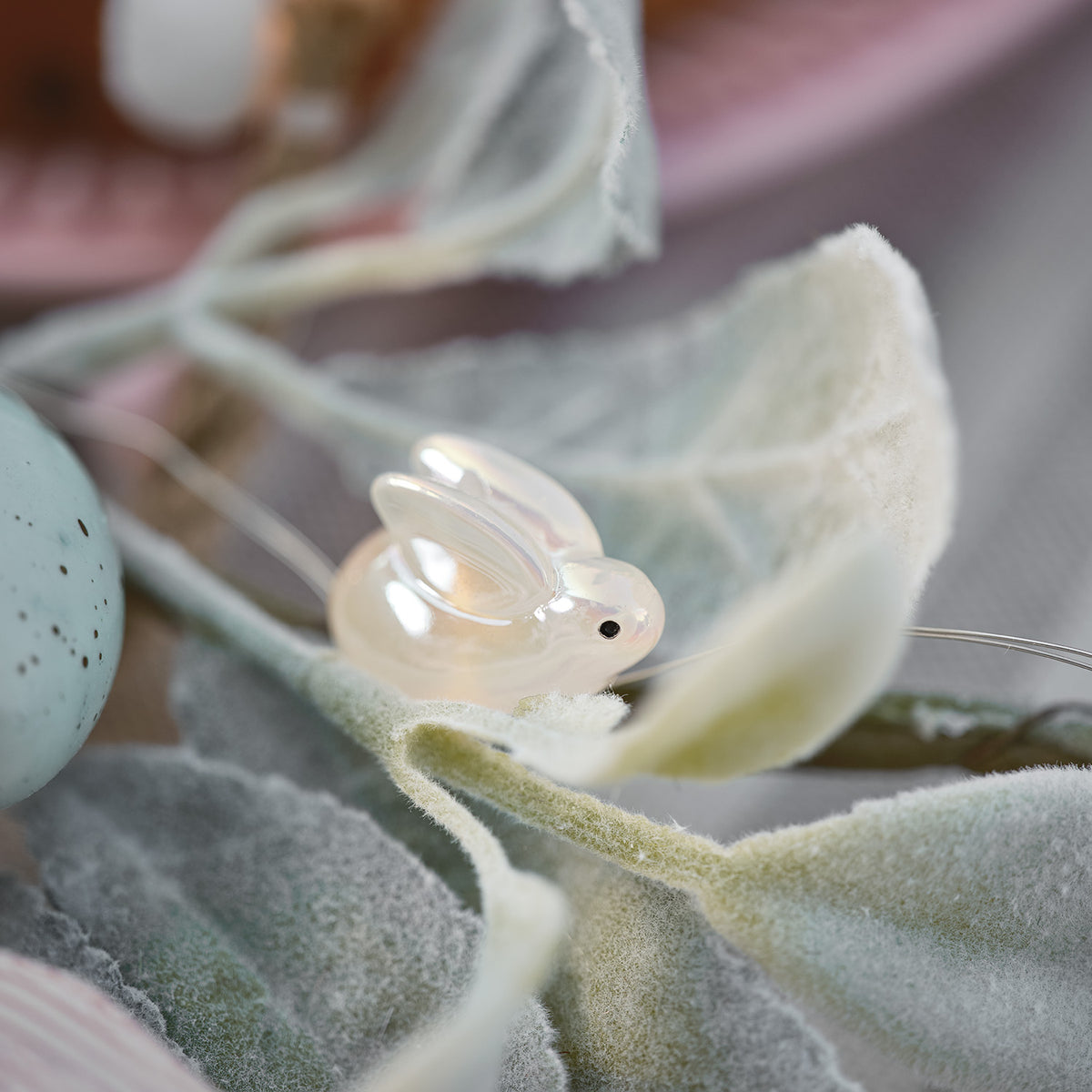 Perlmuttfarbener Hase aus Kunstharz auf samtigem grünen Blatt neben einem blauen Osterei