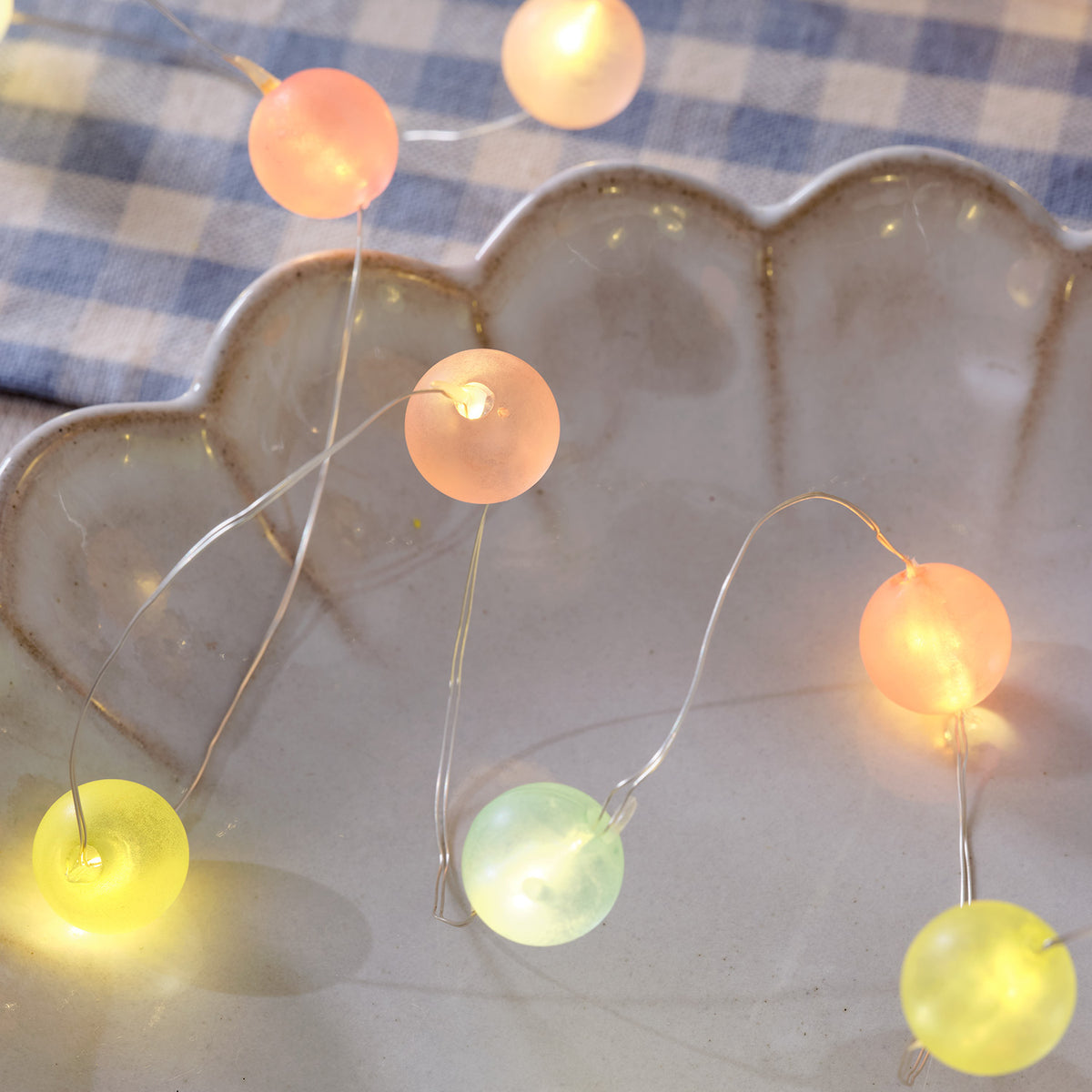 Pastellfarbene LED Kugeln einer Lichterkette liegen auf einer grauen Muschel-Schale auf blau kariertem Stoff