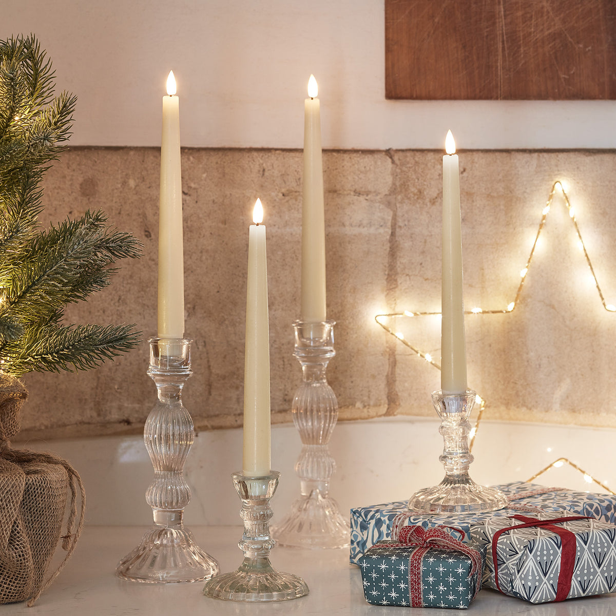 Drei LED Stabkerzen in Glas-Kerzenhaltern neben Weihnachtsgeschenken und Deko