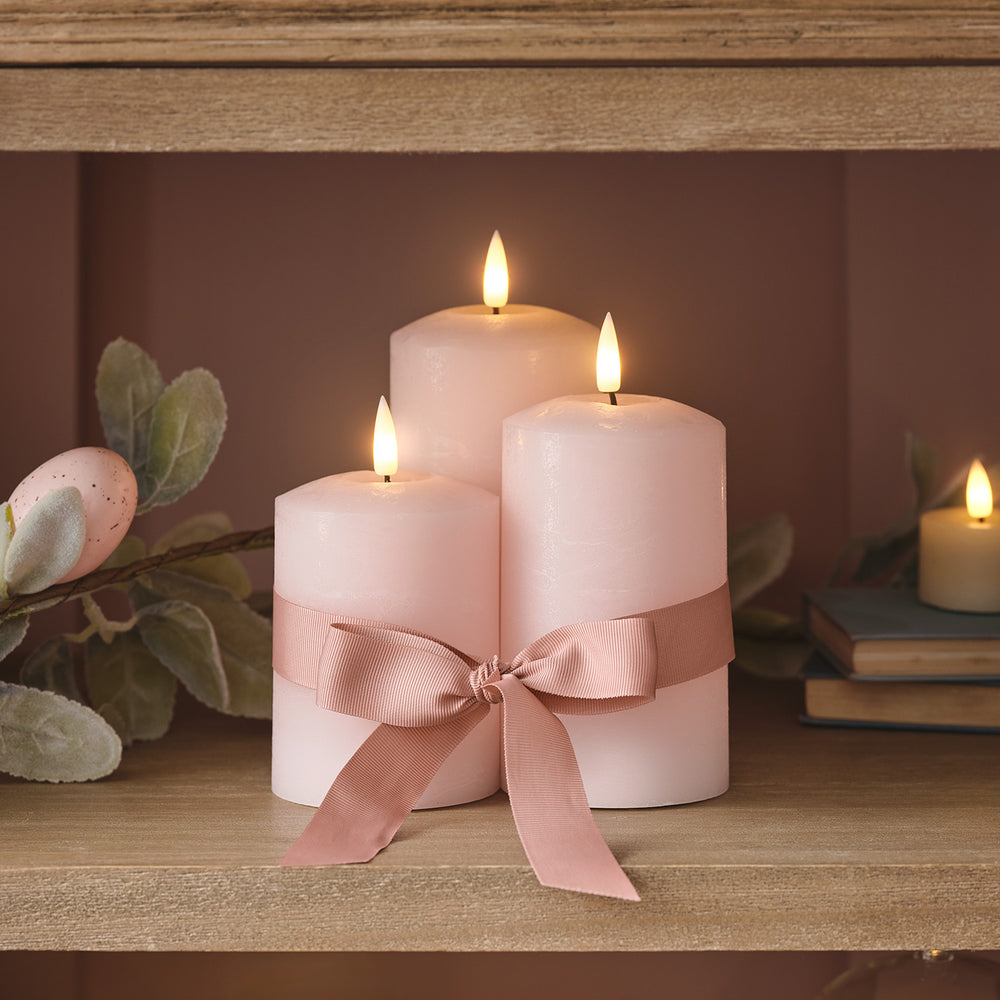 Drei rosa LED Stumpenkerzen mit Schleife auf Holzregal mit Osterdeko
