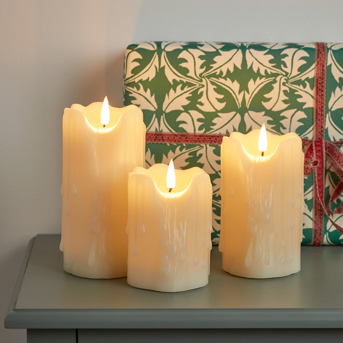 Drei leuchtende LED-Kerzen mit Tropfwachs vor grün-weiß gemustertem Geschenk mit rotem Band