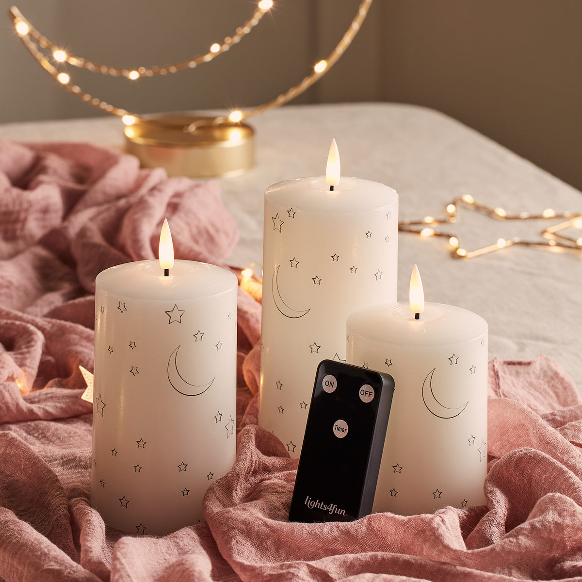 Drei weiße LED-Kerzen mit Mond- und Sternmotiven und Fernbedienung auf rosa Stoff