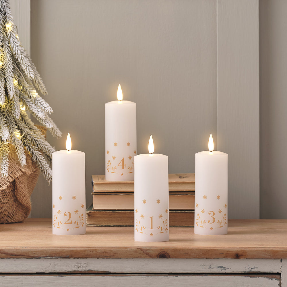 Vier weiße LED Adventskerzen mit goldenen Zahlen 1 bis 4 auf Holztisch neben beleuchtetem Weihnachtsbaum
