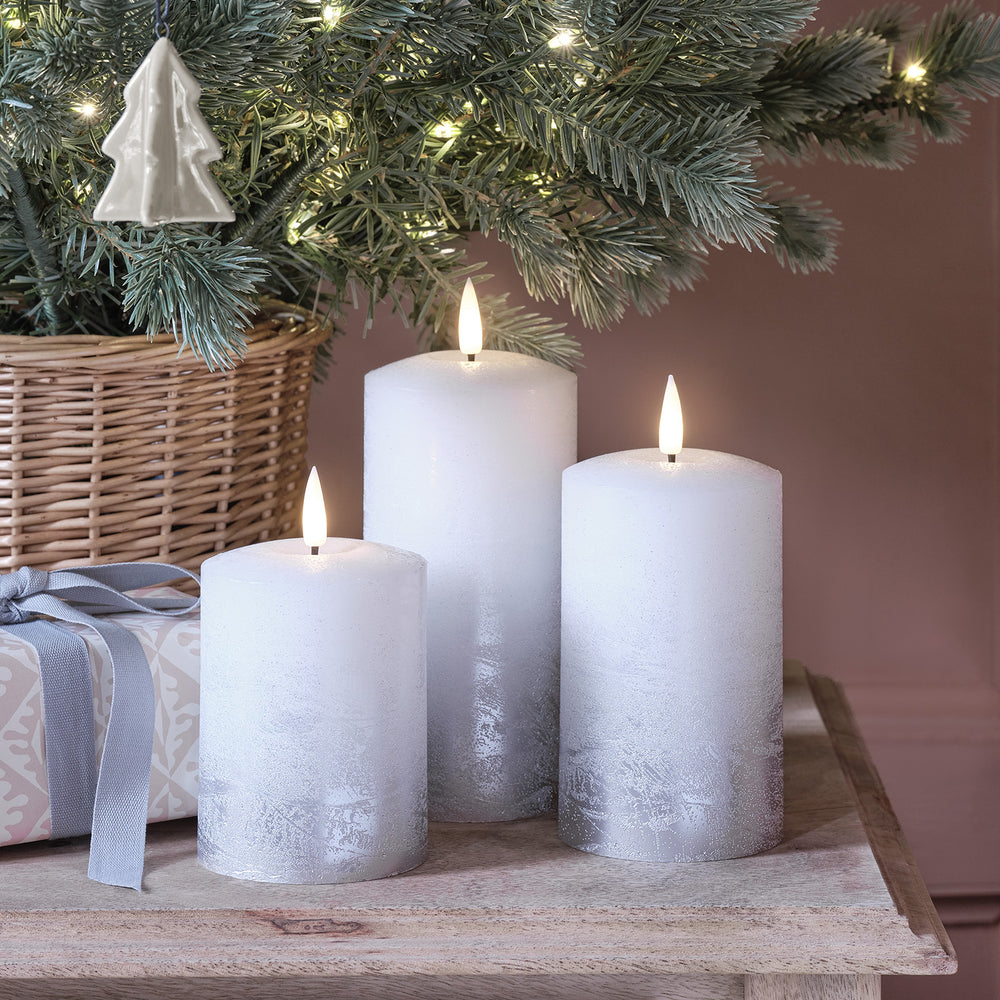Drei silber ombré LED Echtwachs-Kerzen vor einem Weihnachtsbaum mit Geschenk