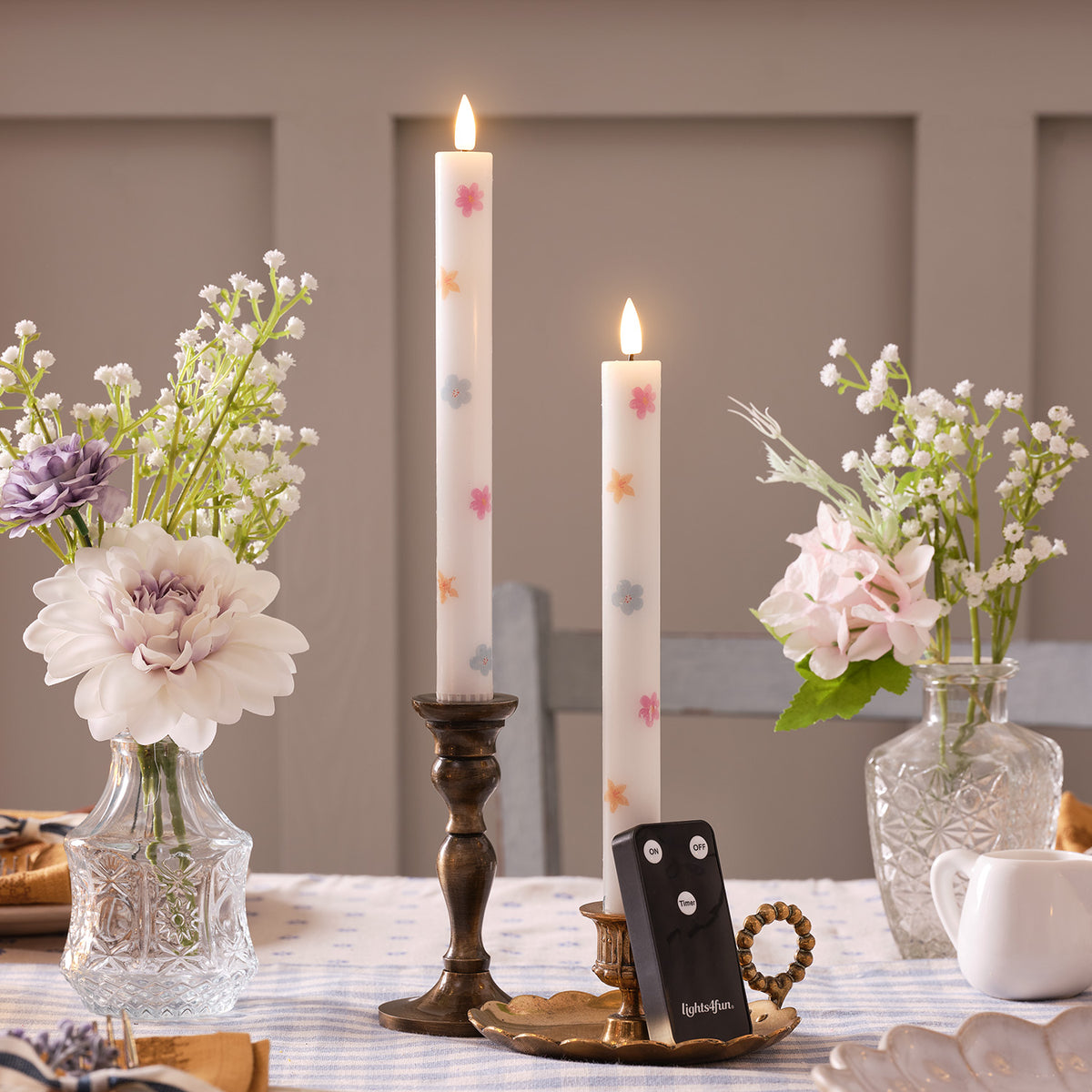Zwei weiße LED Stabkerzen mit buntem Blumenmuster in Kerzenhaltern auf gedecktem Tisch mit Blumenvasen