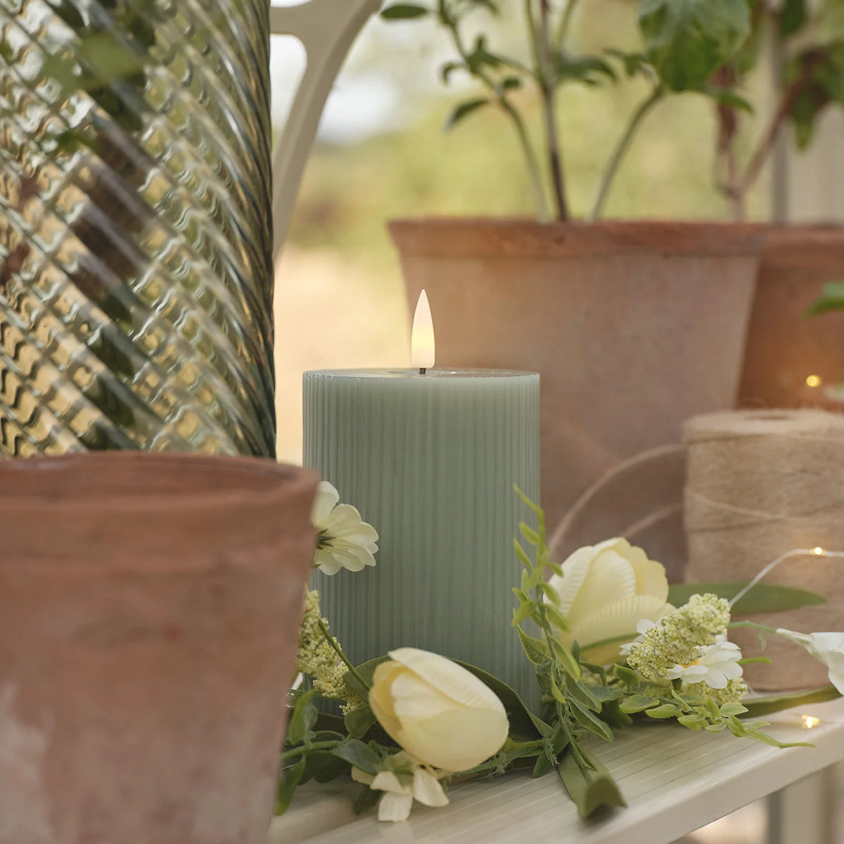 Nahaufnahme einer salbeigrünen LED-Stumpenkerze umgeben von Blumen und Tontöpfen