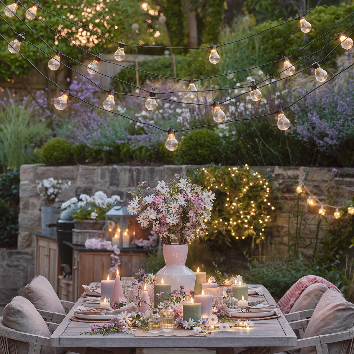 Gedeckter Gartentisch mit salbeigrünen LED-Stumpenkerzen, Blumen und Lichterketten
