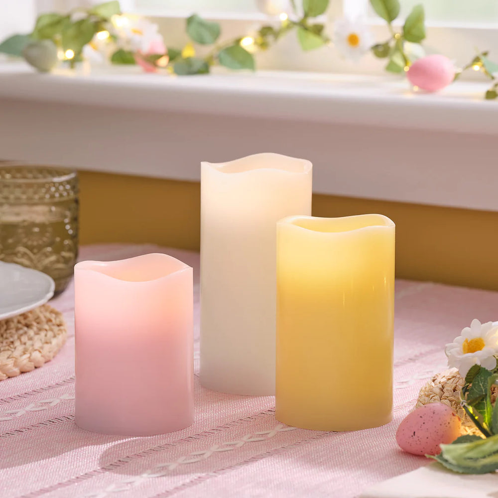Drei LED-Stumpenkerzen in Pastellfarben rosa, weiß und gelb auf rosa Tischdecke mit Frühlingsdeko