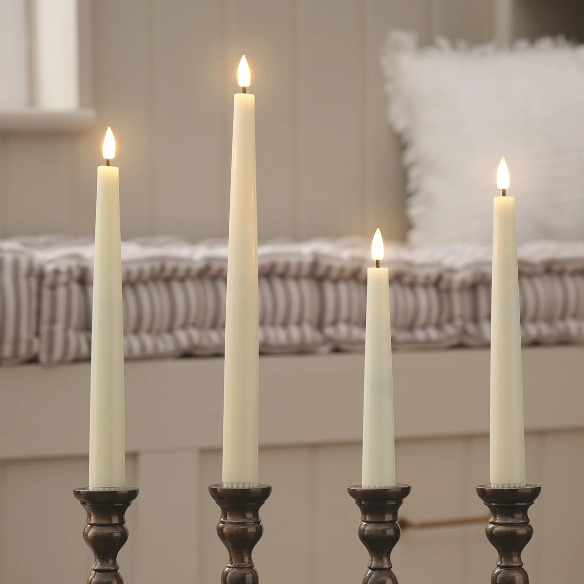Vier LED-Stabkerzen in Metallhaltern mit flackernden Flammen vor einem Bett mit gestreifter Bettwäsche