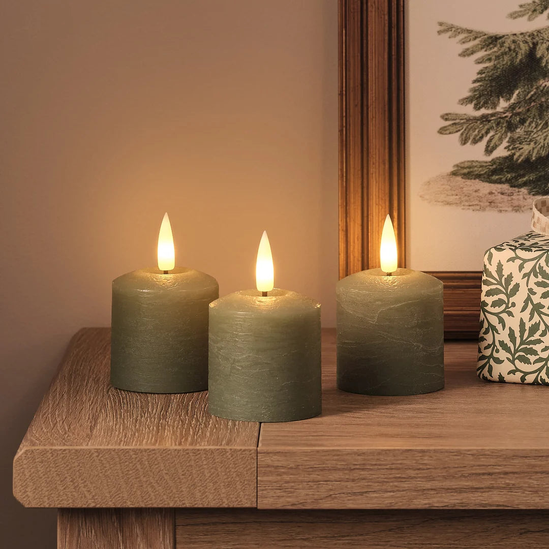 Drei LED-Kerzen in Olivengrün mit weißen LED-Flammen auf massivem Holztisch, neben beige-cremefarbener Leinenserviette mit floralem Muster und Tannendekorations.