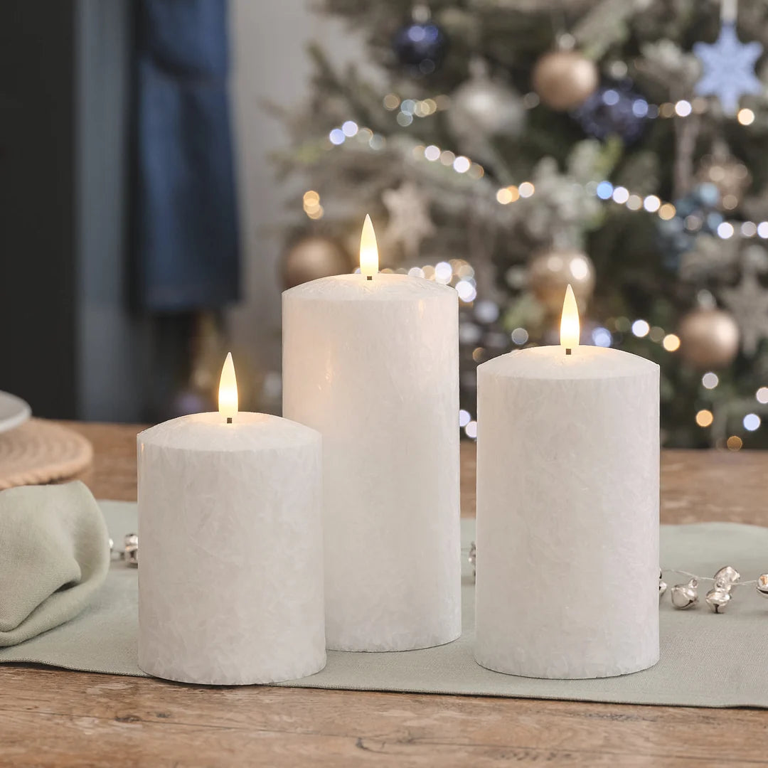 Drei weiße LED-Kerzen in verschiedenen Höhen mit warmweißen LED-Flammen auf Holztisch, beige Leinenserviette, verschwommener Weihnachtsbaum mit Kugeln im Hintergrund.