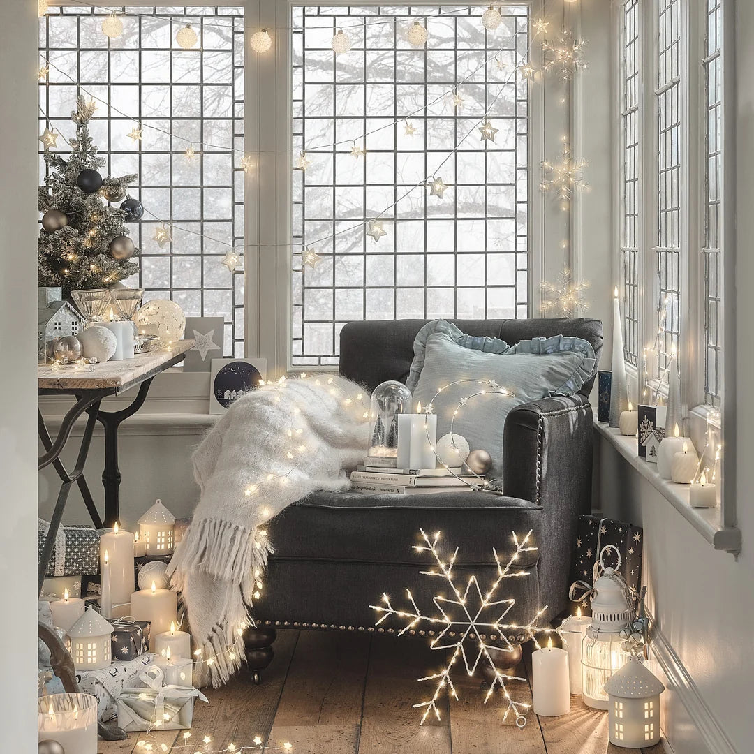 Modernes Wohnzimmer mit schwarzem Sofa, großen Fenstern mit Weihnachtsdeko, weißem Tannengrün mit Kugeln und Schneeflocken-Ornamenten, hängenden LED-Lichterketten, weißen Kerzen, silbernen Rentier-Dekoelementen und weiße LED-Lichterketten.