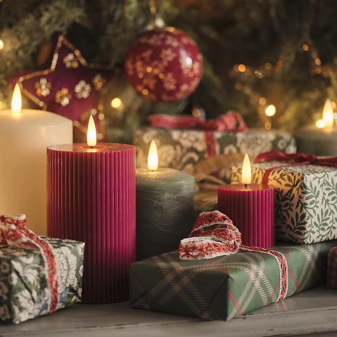 Festliche Weihnachtsdekoration mit roten strukturierten LED-Zylinderkerzen mit warmweißen LED-Flammen, cremefarbenen Zylinderkerzen, grauem Kerzenständer, Geschenkboxen in Rot und Grün mit roten Schleifen, grüne Girlande, Weihnachtskugeln in Rot und Beige, Holztisch.