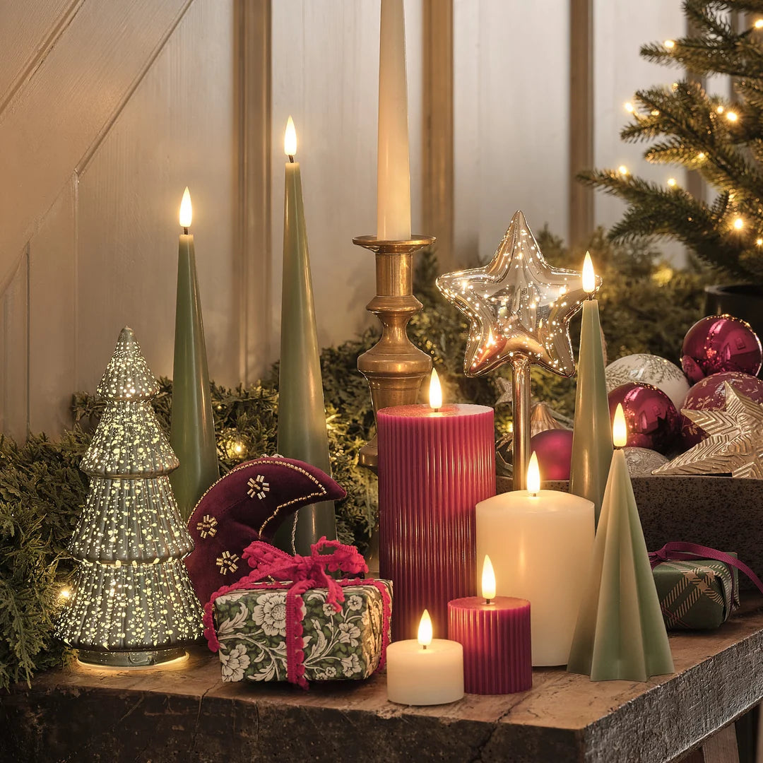 Kamindekoration mit roten strukturierten LED-Zylinderkerzen, cremefarbenen LED-Zylinderkerzen, cremefarbenen Taper-Kerzen auf Kerzenständer, goldenes Kerzenständer-Set und grüne Girlande auf Holzoberfläche.