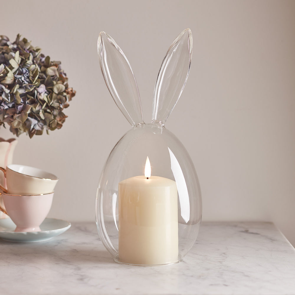 Glasglocke mit Hasenohren und brennender TruGlow Kerze auf Marmorplatte neben Teetassen und getrockneter Blume