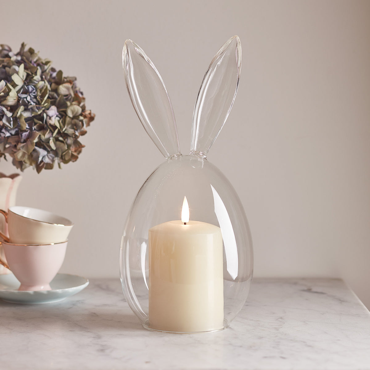 Glasglocke mit Hasenohren und brennender TruGlow Kerze auf Marmorplatte neben Teetassen und getrockneter Blume