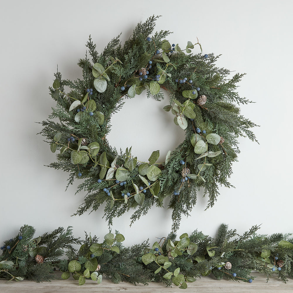 Weihnachtskranz ohne Beleuchtung mit grünen Tannenzweigen, Eukalyptusblättern, blauen Beeren und Tannenzapfen an weißer Wand