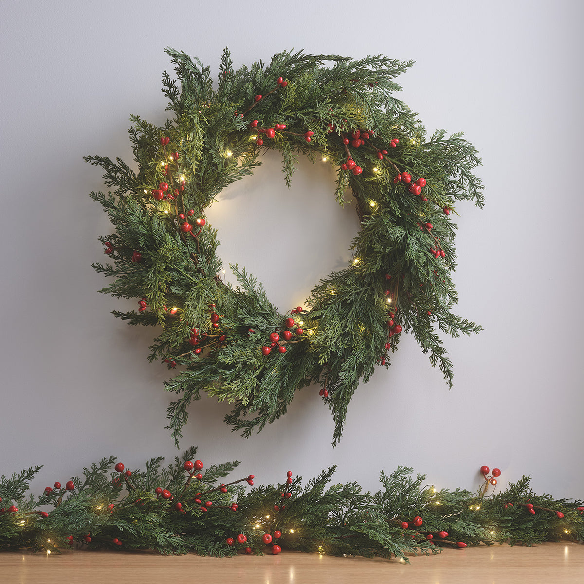 Weihnachtskranz mit roten Beeren und warmweißen LED-Lichtern vor weißer Wand mit Girlande