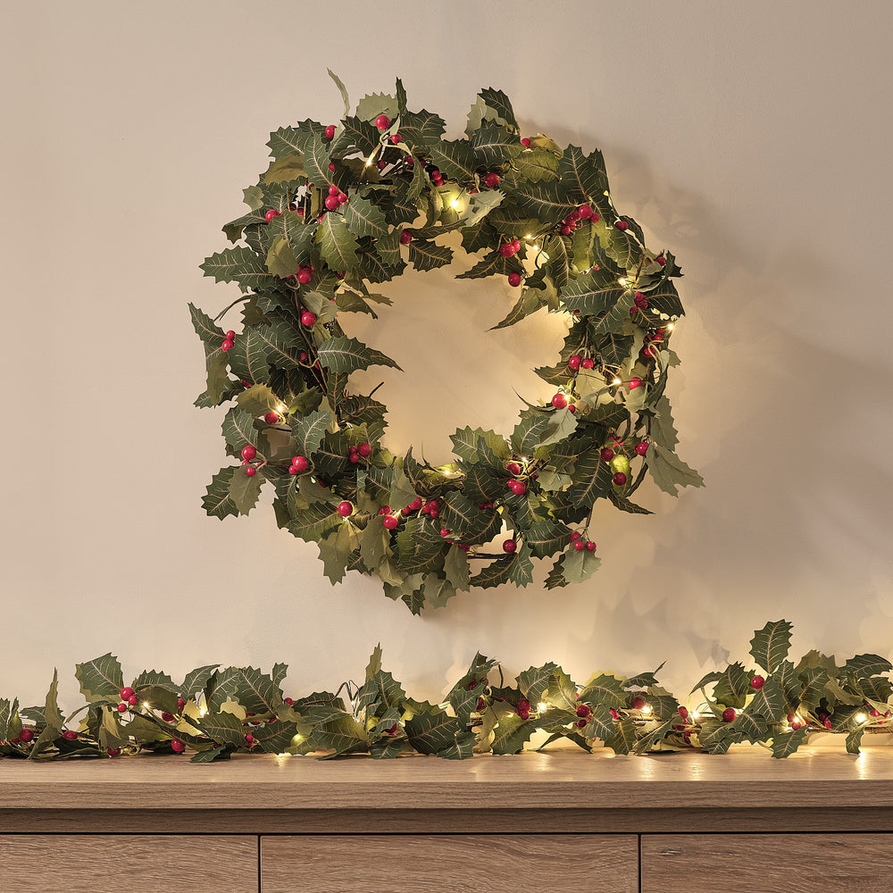 Beleuchteter Weihnachtskranz mit Stechpalmenblättern und roten Beeren an Wand über Sideboard