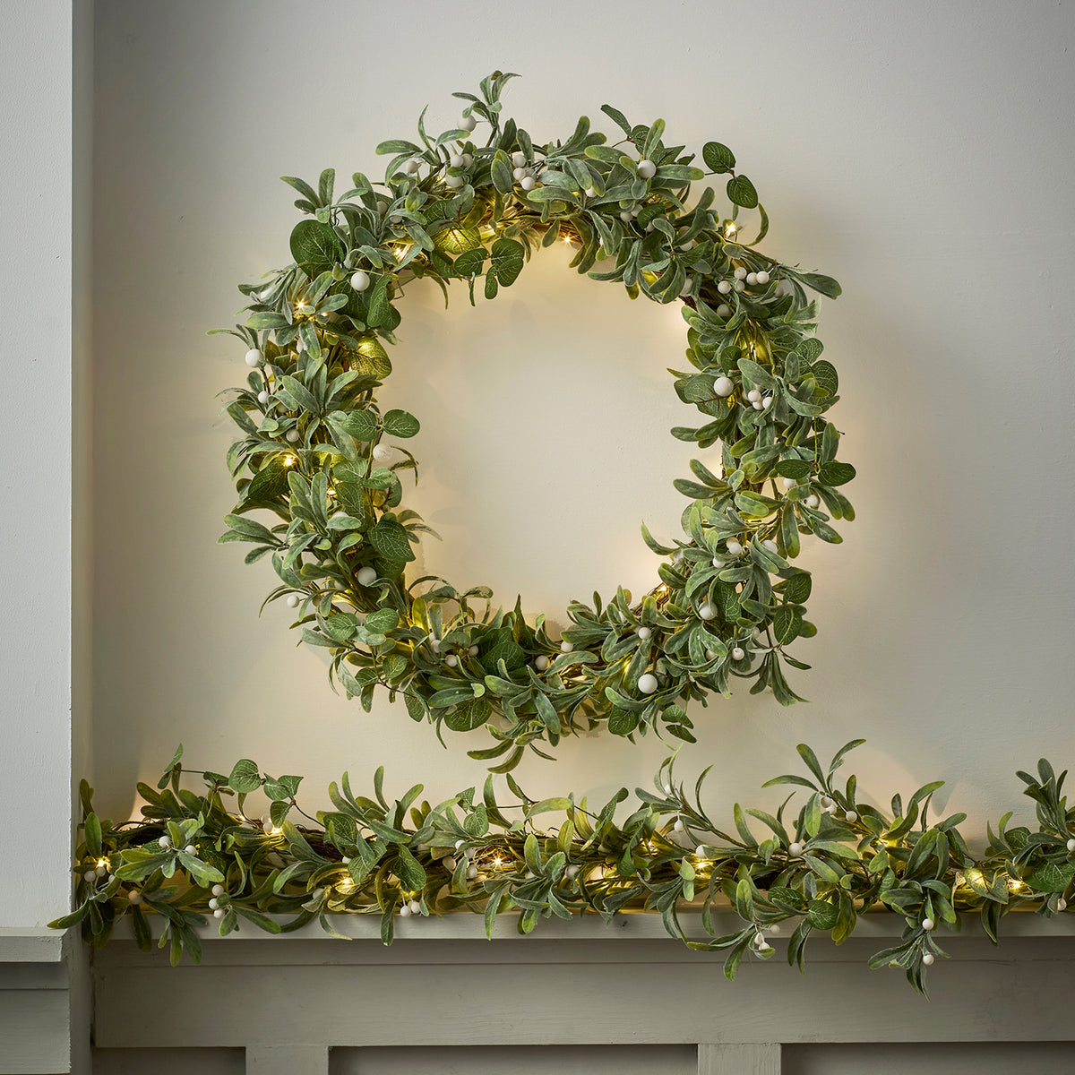 Beleuchteter Weihnachtskranz mit grünen Blättern und weißen Beeren an weißer Wand