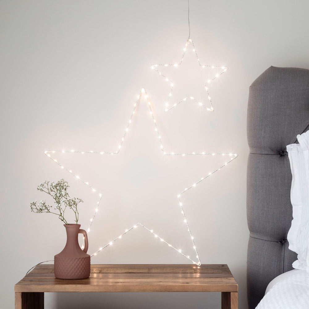 Zwei weiße LED-Drahtsterne in verschiedenen Größen an einer Wand neben einem grauen Bett