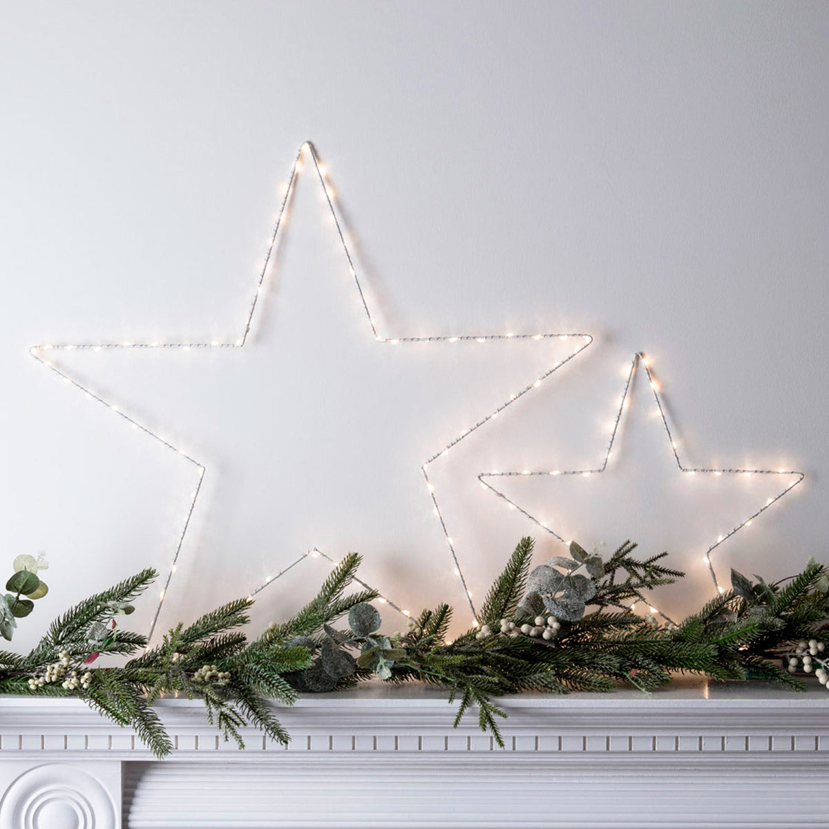 Zwei LED-Drahtsterne an weißer Wand mit Tannenzweigen und Weihnachtsdeko auf Kaminsims