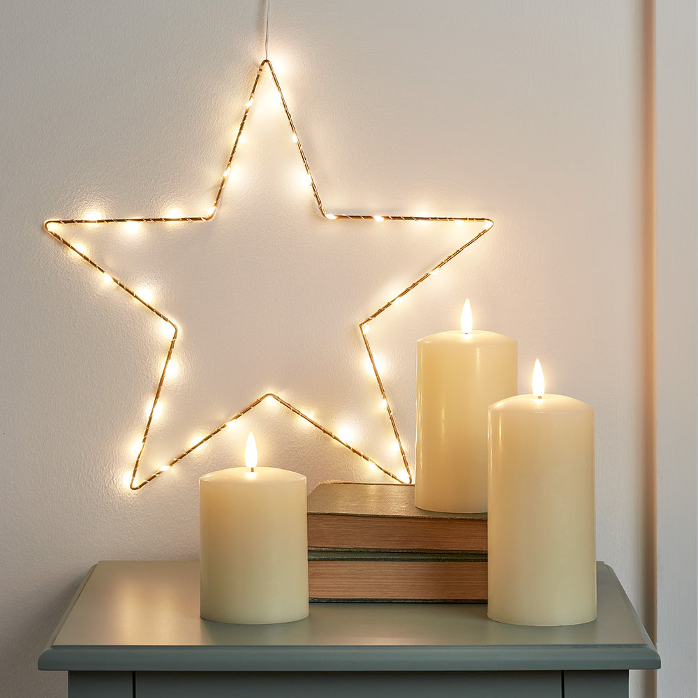 Goldener Stern mit warmweißen LEDs an Wand und drei elfenbeinfarbene TruGlow® Stumpenkerzen auf grauem Tisch
