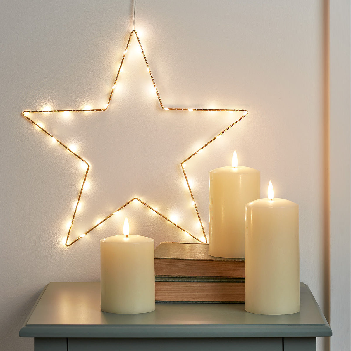 Goldener Stern mit warmweißen LEDs an Wand und drei elfenbeinfarbene TruGlow® Stumpenkerzen auf grauem Tisch