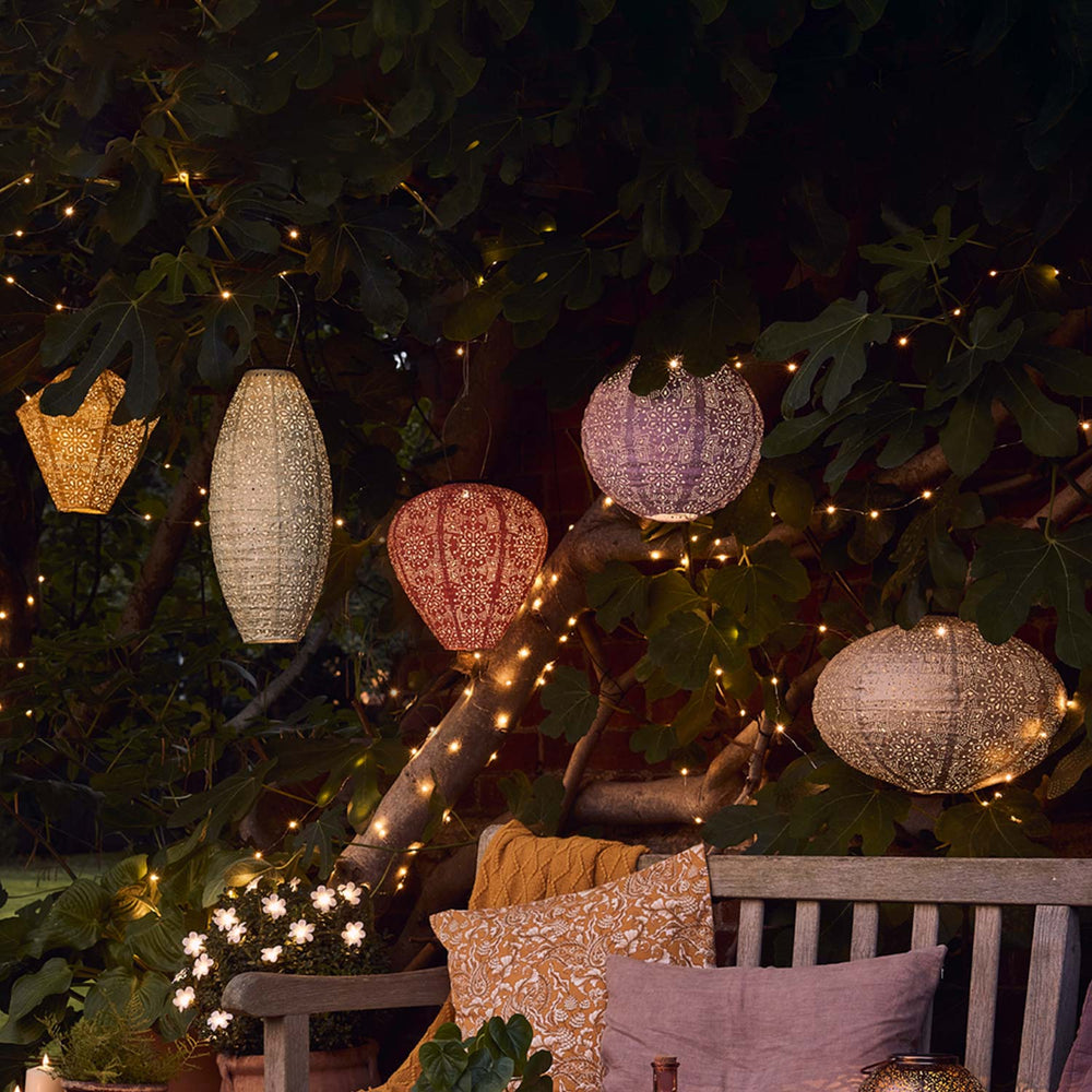 Fünf bunte Tyvek Solar Lampions mit Blumenmuster hängen beleuchtet an einem Baum über einer Holzbank.