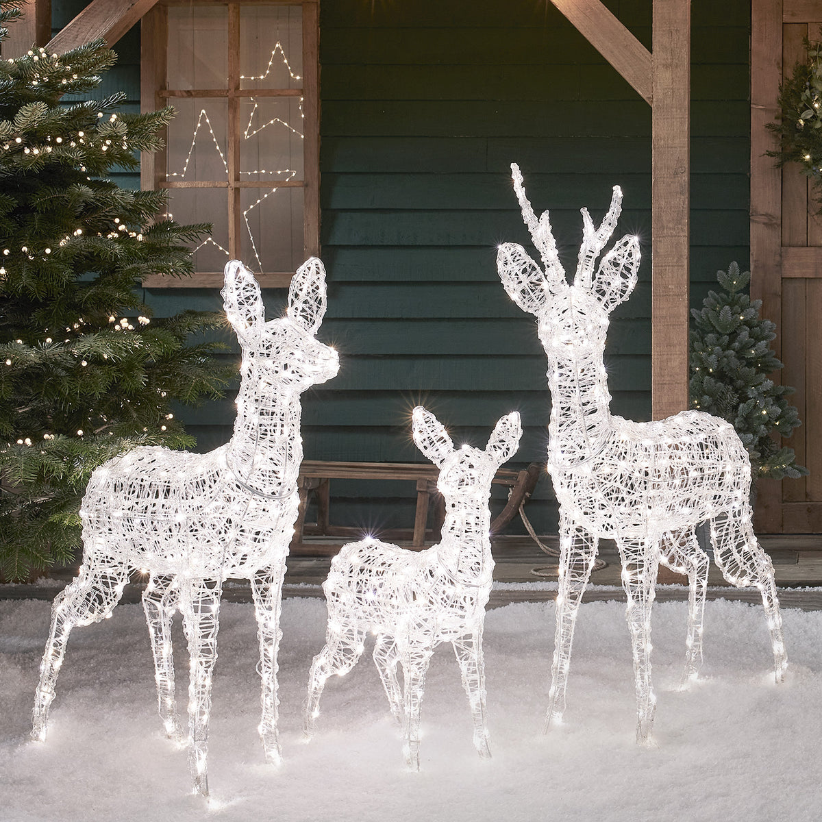 Drei weiße LED-Rentierfiguren aus Acryl auf Schnee vor grünem Holzhaus mit Fensterdeko