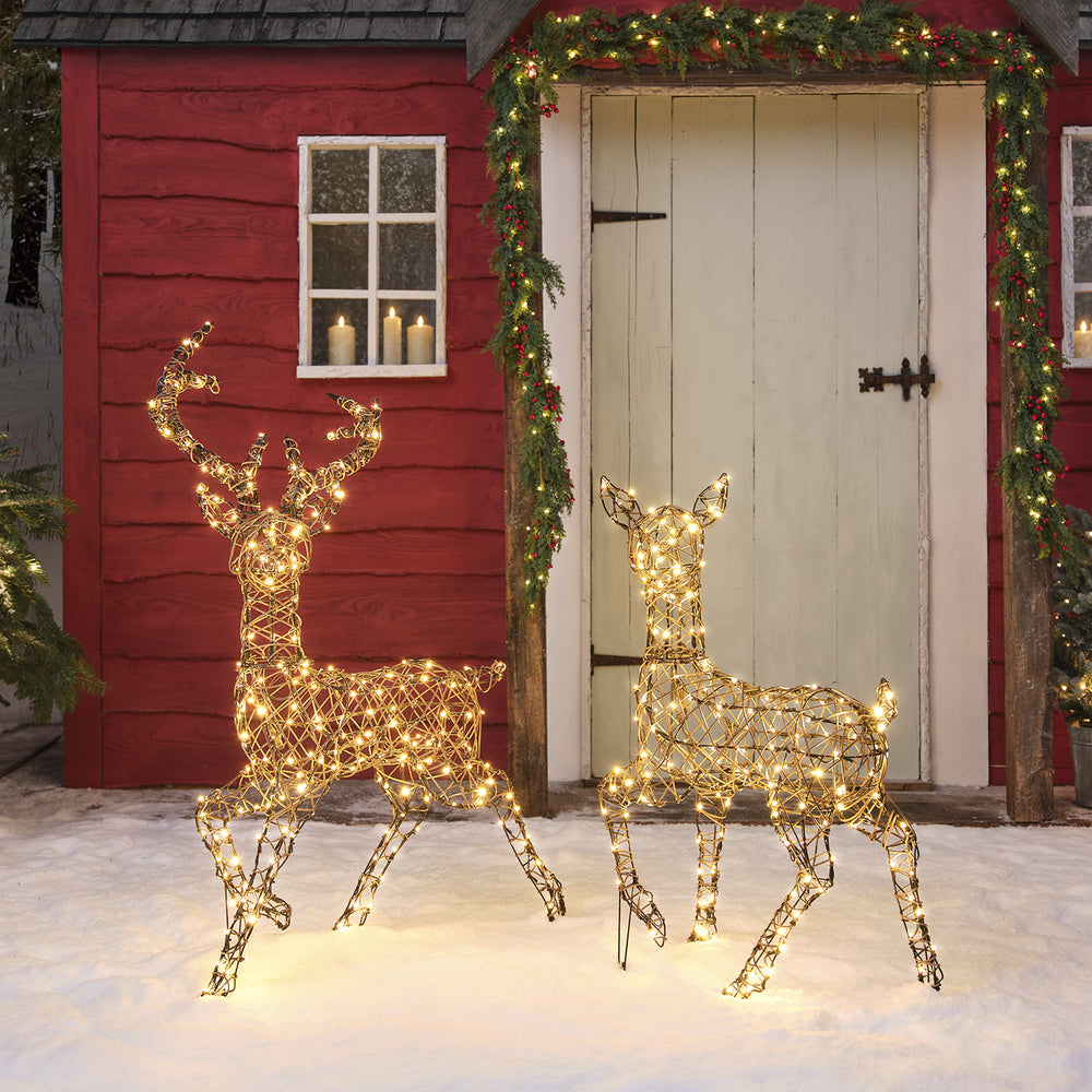 LED-Rentier Hirsch und Hirschkuh aus Kunst-Rattan mit warmweißen Lichtern vor rotem Haus im Schnee