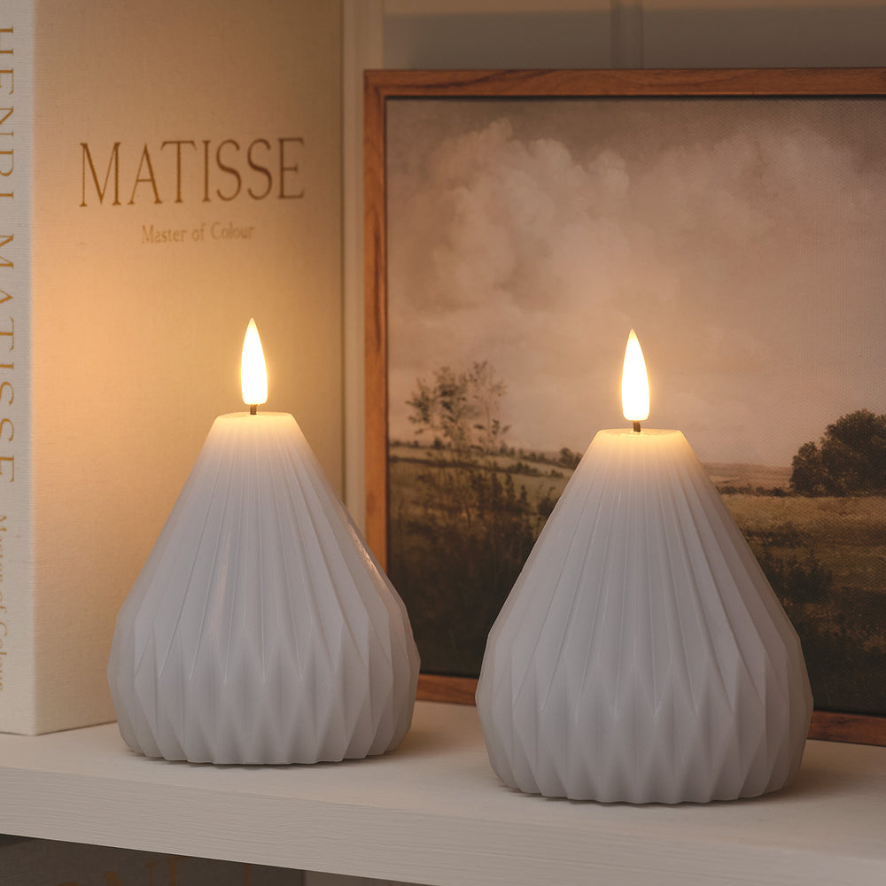 Zwei hellgraue LED-Echtwachskerzen im Pear-Design mit warmweiß flackernder Flamme auf Regal vor Bildern
