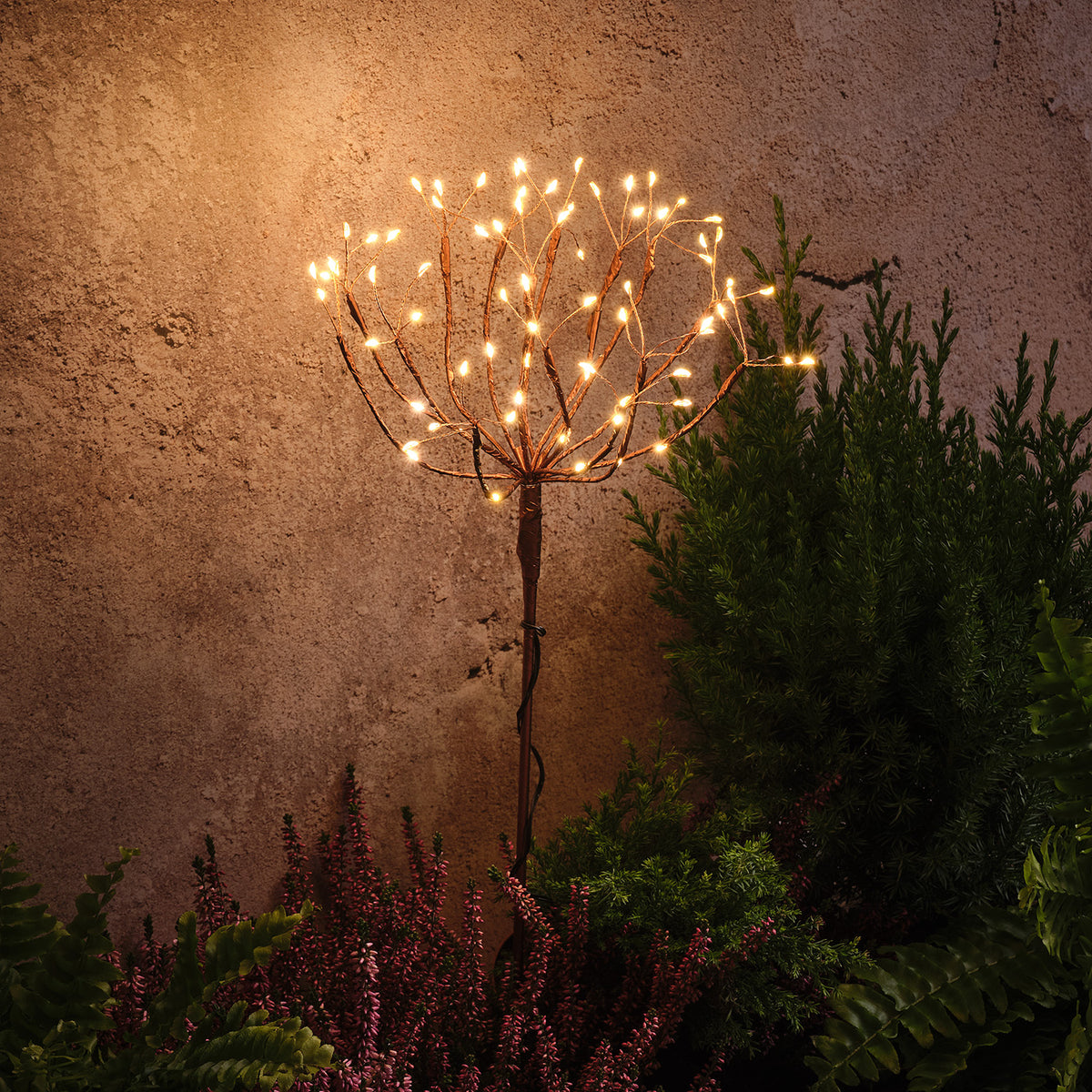 Einzelner goldfarbener Solar Feuerwerk Gartenstecker mit leuchtenden warmweißen LEDs vor Wand