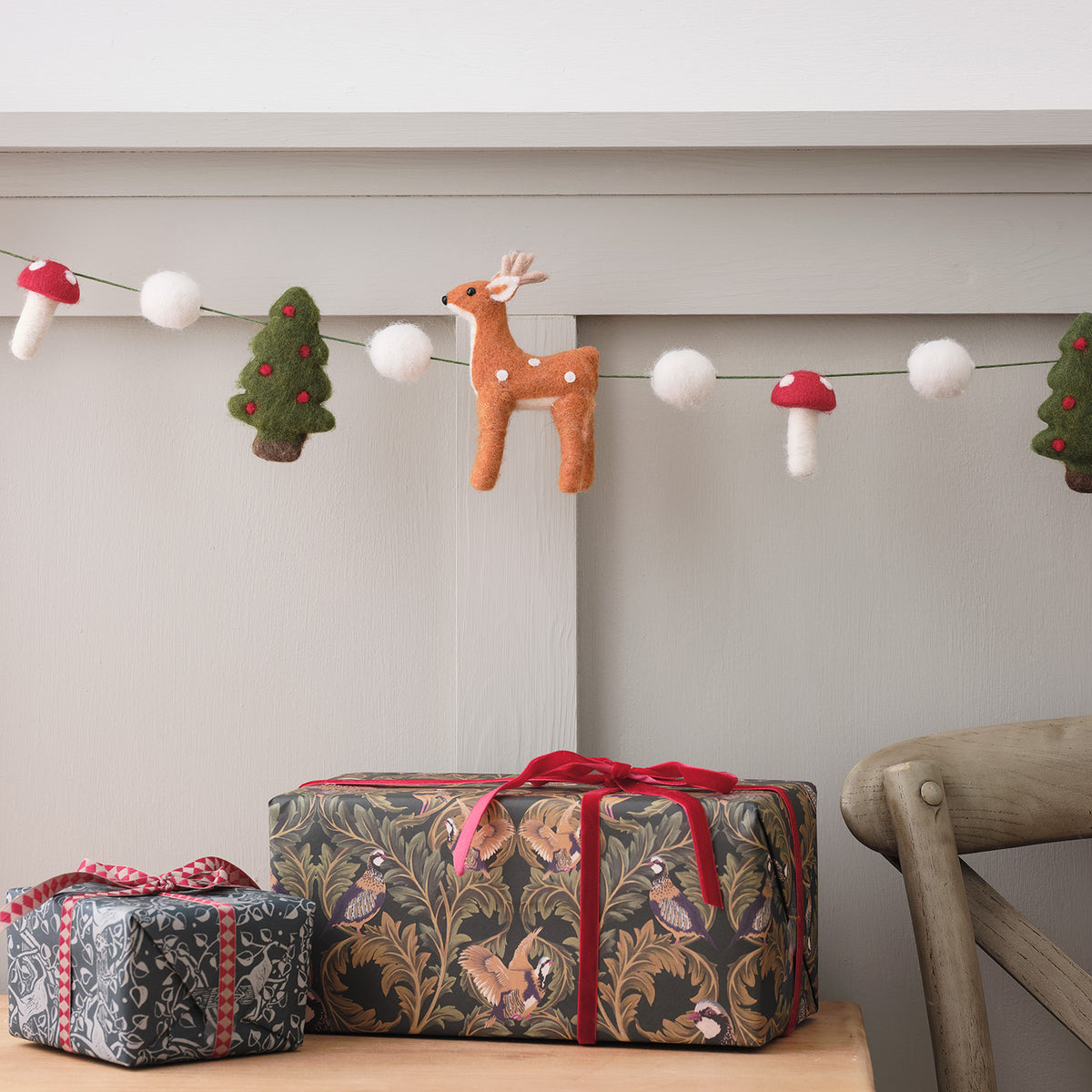 Weihnachtsgirlande aus Filz mit Rentier, Tannenbäumen, Pilzen und weißen Kugeln über einem Tisch mit Geschenkpaketen
