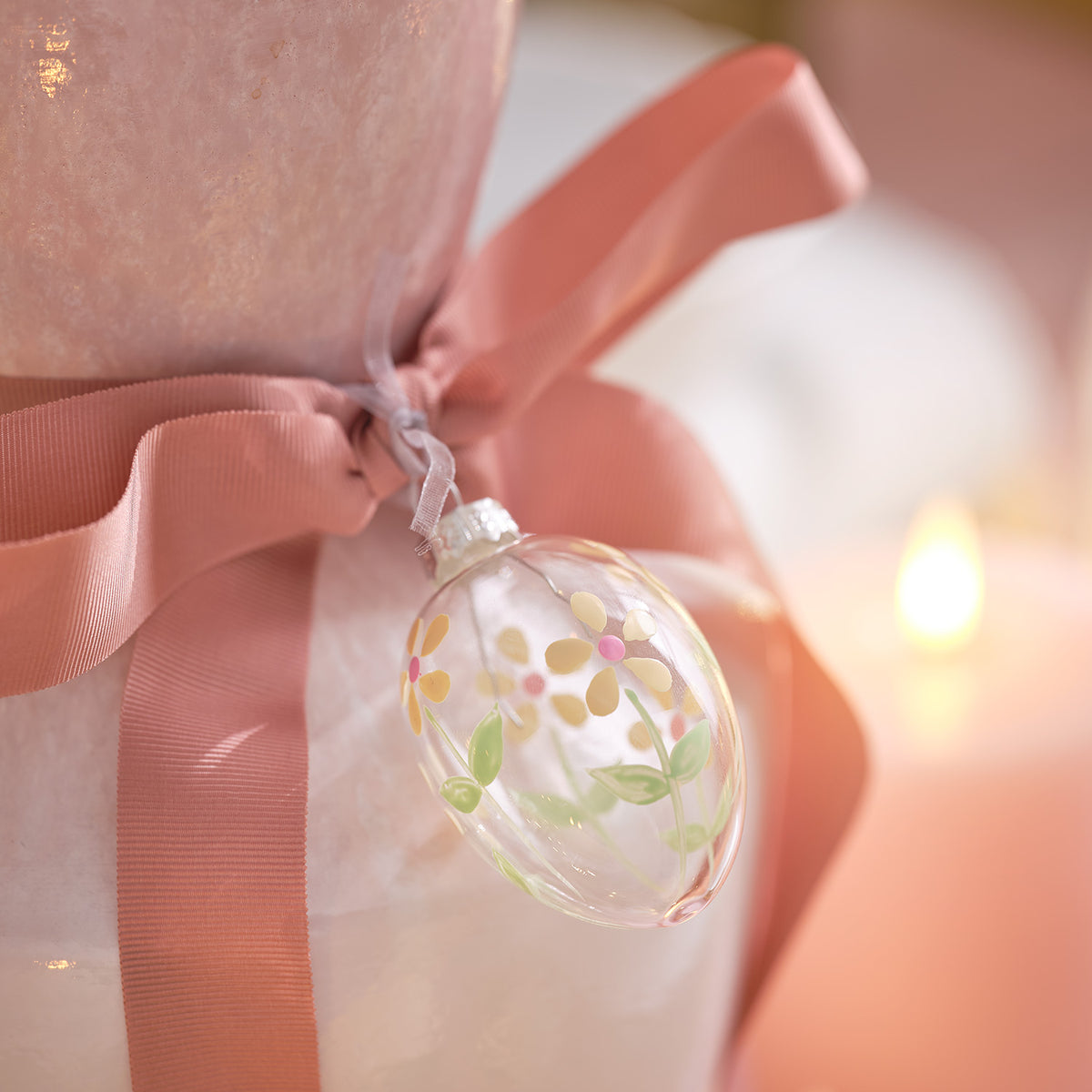 Nahaufnahme eines Glas-Ostereis mit Blumenmotiv an rosa Schleife vor Kerzenlicht