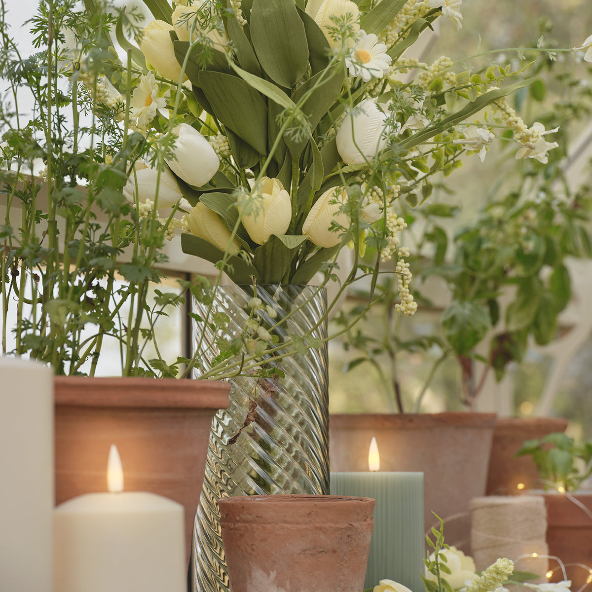Grüne Glasvase mit Struktur gefüllt mit Tulpen, umgeben von Terrakottatöpfen und Kerzen im Gartenhaus