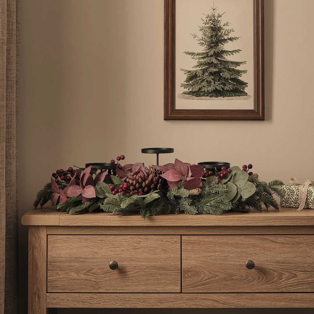 Stimmungsvolles Adventsgesteck auf Holzkommode mit drei burgundy-roten strukturierten LED-Taperkerzen in schwarzen Haltern, umrahmt von grünem Fichtengrün, burgundy Blattwerk, roten Beeren, Zapfen und eleganten grün-cremefarbenen Dekoelementen – edle Weihnachtsdekoration mit gerahmtem Tannenbaum-Kunstdruck im Hintergrund für festliche Wohnkultur.