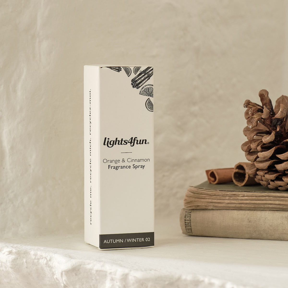 Elegantes lights4fun Raumduft-Spray in cremefarbener Verpackung mit botanischem Blattmotiv, präsentiert neben gestapelten Leinenbüchern, braunen Pinienzapfen und natürlichen Holz-Akzenten auf neutrale Leinenhintergrund – hochwertiges Duftprodukt für stilvolle Raumbeduftung und festliche Wohnzimmer-Dekoration mit Premium-Verpackungsdesign