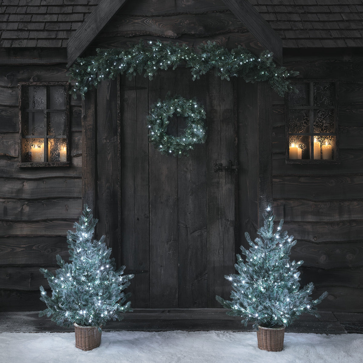 Weihnachtskranz, Girlande und zwei Weihnachtsbäume mit kaltweiß leuchtenden LEDs vor Holztür