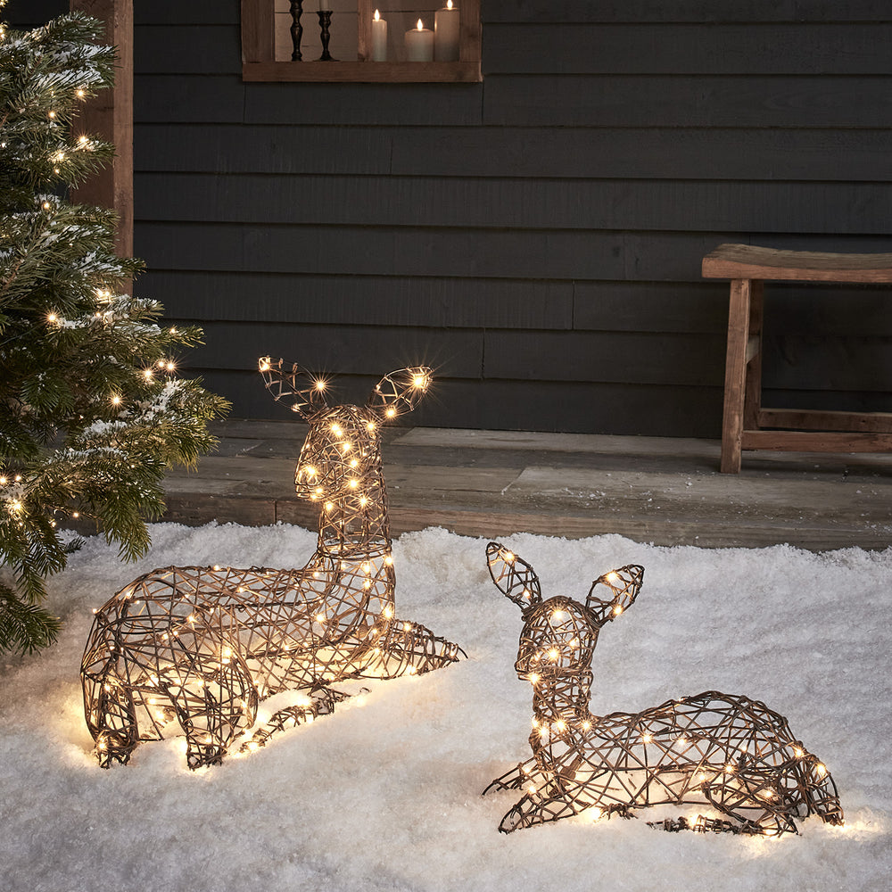 Liegende LED-Rentiere Mutter und Kind aus braunem Rattan mit warmweißen Lichtern im Schnee vor dunkler Wand