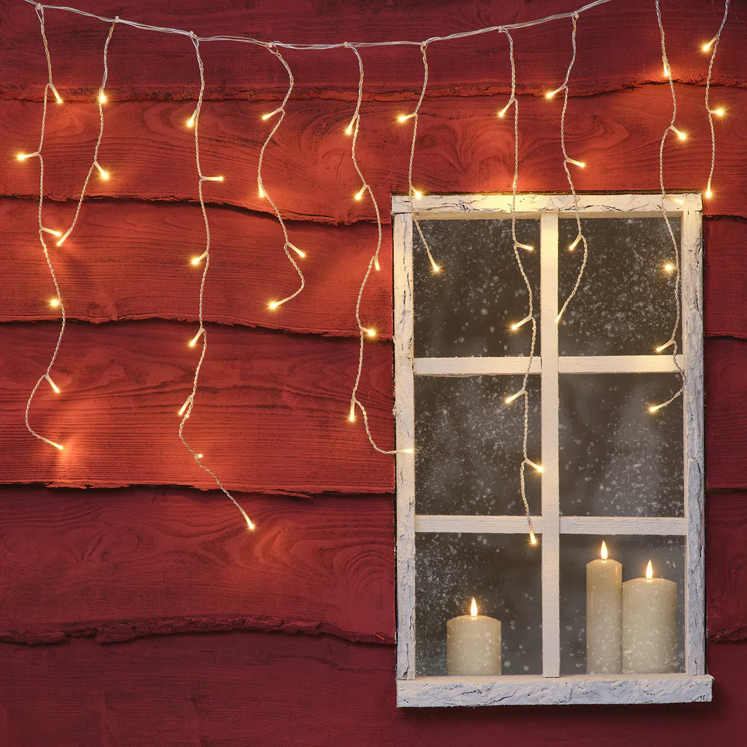 Warmweiße Eiszapfen-Lichterkette mit 100 LEDs hängt vor roter Holzfassade neben Fenster mit brennenden Kerzen
