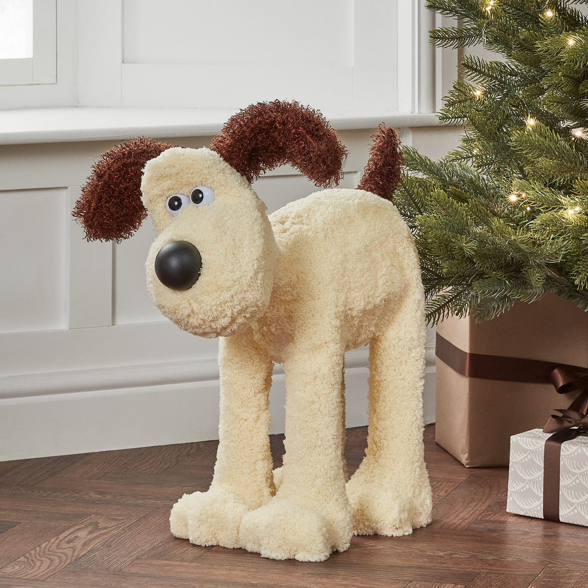 Gromit LED-Figur mit rot-weißem Schal auf Holzuntergrund vor Weihnachtsbaum und Geschenken