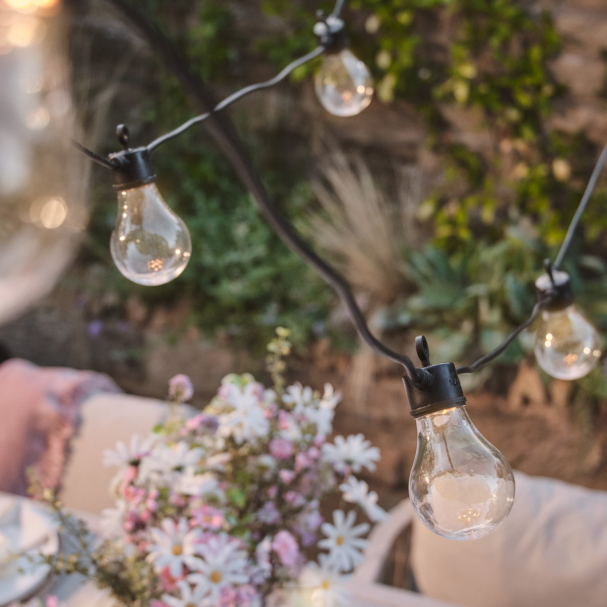 Nahaufnahme einer warmweißen Glühbirne der Lichterkette vor Blumen und Sitzbereich