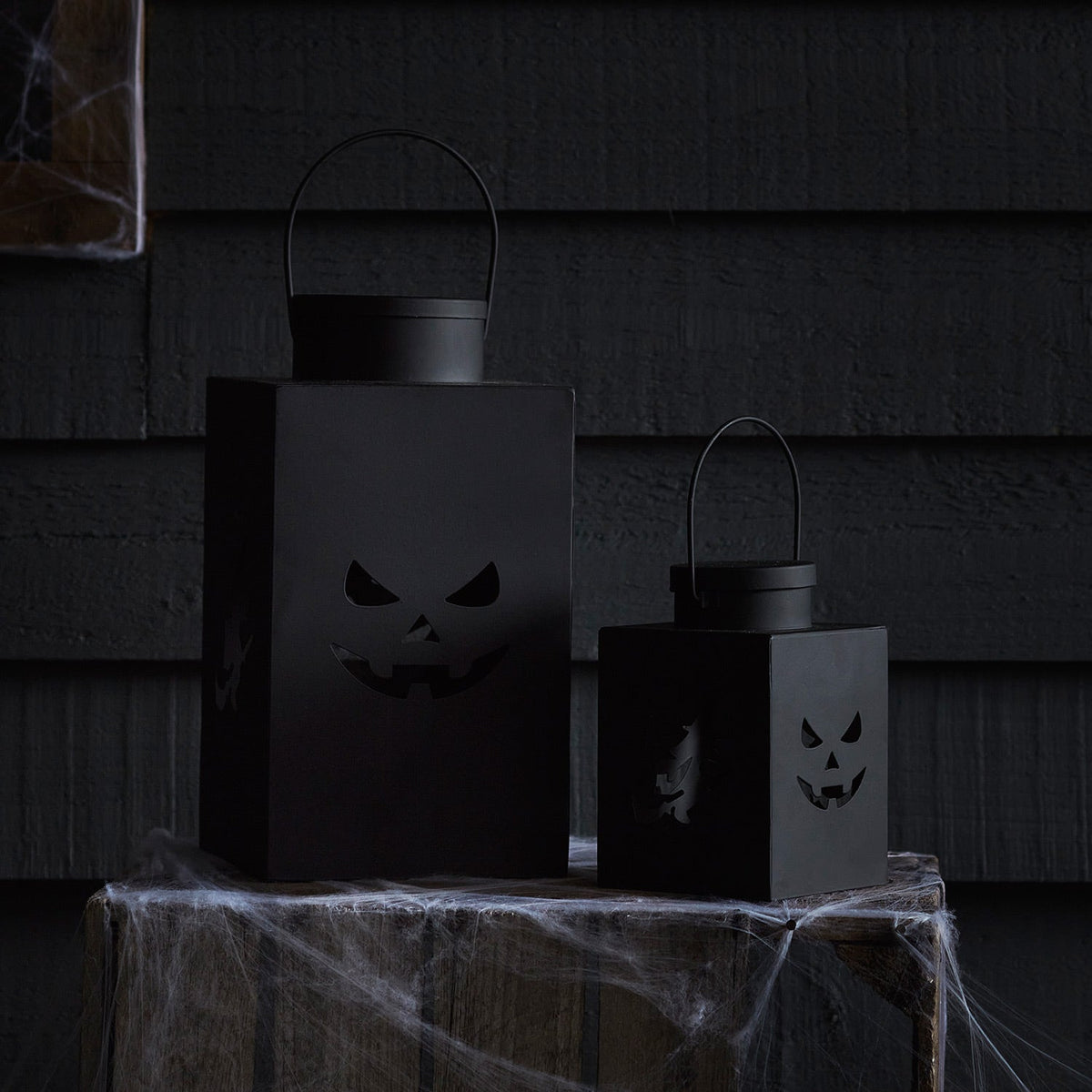 Zwei schwarze Metalllaternen mit Halloween-Motiven auf Holztisch mit Spinnweben vor dunkler Wand
