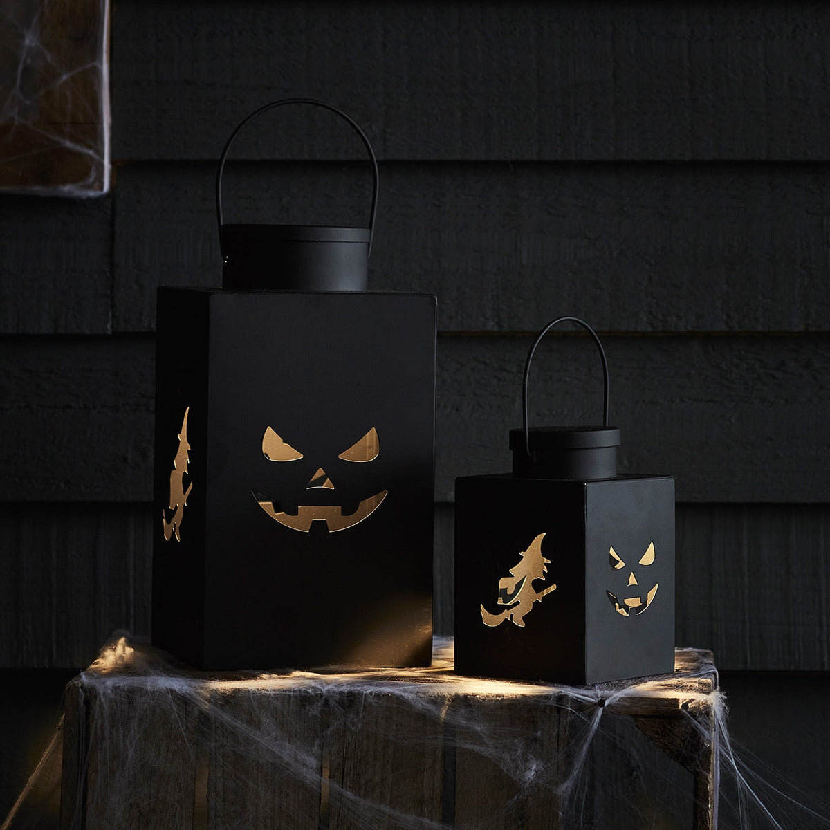 Halloween-Laternen aus schwarzem Metall mit Hexen- und Kürbis-Motiven, beleuchtet auf Holztisch mit Spinnweben